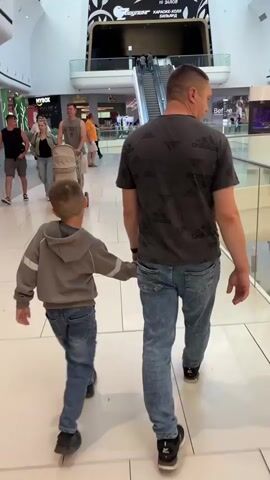 A father and son walk hand-in-hand through a brightly lit shopping mall. The father, wearing a dark t-shirt with a repeating logo pattern and blue jeans, is on the right. The son, in a grey hooded jacket and blue jeans, is on the left. They are both walking away from the camera. In the background, other shoppers can be seen, including a woman pushing a stroller and a man in an orange shirt near an escalator. The mall features a modern design with multiple levels, escalators, and storefronts.