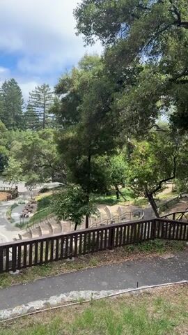 The video pans across a park-like setting with lush green trees and a cloudy sky. In the foreground, a dark brown wooden fence runs horizontally across the frame, with a paved pathway behind it. Beyond the fence, a curved amphitheater-style seating area is visible, with many steps leading down. Further into the park, there's a playground area with sand and some wooden structures. A white portable toilet is also visible near the playground. A single person, possibly a child, is seen walking on a paved path in the playground area. The overall environment appears serene and natural.