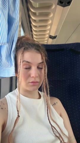 A woman sits on what appears to be a train, her hair in a messy bun with some strands braided. She wears a white tank top and faces the camera. Initially, she looks serious, then playfully makes a kissing face before flashing a peace sign. Tattoos are visible on her arms. The train interior features a blue-striped curtain and seats with a patterned fabric. Overhead are air conditioning vents. The video appears to be shot handheld.