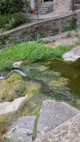 A moving body of water cascades over rocks in a natural setting. Green vegetation surrounds the water. The camera perspective is from above and slightly to the side, looking down at the scene. There are no people visible in the video.