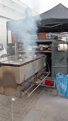 A man wearing a black baseball cap and a red apron is standing next to a large, multi-compartment smoker that is emitting a lot of smoke. The smoker is attached to a trailer and appears to be set up outdoors, possibly in a parking lot, as indicated by the paved ground and a building with a "TEDI" sign in the background. The man is wearing a red protective glove on his right hand and is reaching towards the smoker, possibly to adjust it or check on the food. The scene suggests food preparation or cooking is in progress.