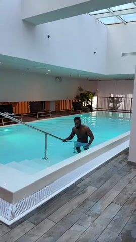 A man with a muscular physique, dark skin, and a beard is seen exiting an indoor swimming pool. He is wearing light blue swim shorts and uses a metal handrail and white steps to slowly ascend from the clear, light blue water. As he walks out, he looks directly towards the camera.

The pool area is brightly illuminated, with natural light filtering in from a skylight overhead. The pool itself is rectangular with pristine white coping and steps. The flooring surrounding the pool features a modern wooden-style design. In the background, behind the pool, several lounge chairs with orange and white striped cushions are visible, along with some green plants, against a clean white wall. The setting appears to be a contemporary and well-maintained indoor pool, likely within a hotel or spa environment. There is no visible text in the video.