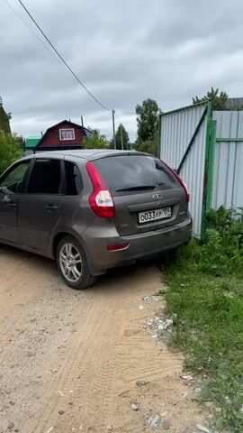 A grey sedan is parked on a dirt path next to a grassy area and a metal fence. A man is sitting in the driver's seat with his hands on the steering wheel. The car is facing towards the right side of the frame. In the background, there are houses and trees under a cloudy sky. The camera is stationary and capturing the scene from the side of the car.