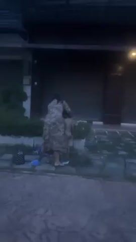 A woman in a patterned dress is standing next to a child, who appears to be naked from the waist up, in front of a building. The woman seems to be adjusting or touching the child's upper back area. A small bush or plant sits between them and the building. The building has dark garage doors and some signage in a non-English script above. There are also some items on the ground near the woman's feet. The scene appears to be taking place outdoors on a paved area, likely in the evening or early morning due to the dim lighting.