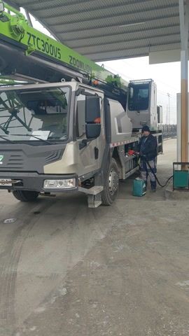 A man is refueling a large green and gray Zoomlion ZTC300V5 crane truck at a gas station. The man is wearing a gray jacket, gray pants, and a dark hat. He is standing near the middle of the truck, holding a fuel nozzle. The truck is parked under a gas station awning, with another large vehicle visible behind it. The ground is concrete.