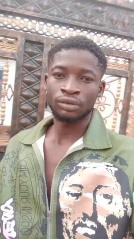 A young black man with a short beard and dark hair, wearing a green jacket with a portrait of a person and text, is walking and filming himself with a mobile phone. He is outside, near a metal fence and other buildings. The video is shaky due to his movement.