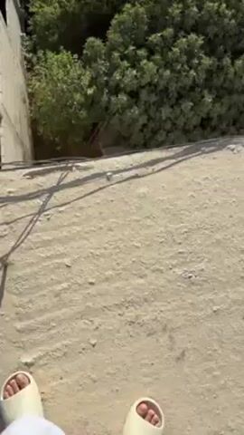 A person stands on a flat roof, looking down into a courtyard. The courtyard is filled with trees and a few buildings. There are two water tanks and what appears to be a chicken coop. The person is wearing white pants and beige sandals.  Loud vehicle noises are audible.