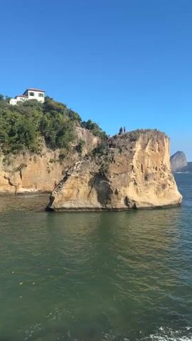 A group of people are gathered on a rocky cliff overlooking the ocean. One person is standing on a ledge near the water, while others are higher up on the cliff face, some appearing to be climbing. The sun is shining, and the sky is clear blue. The water is a greenish-blue color.