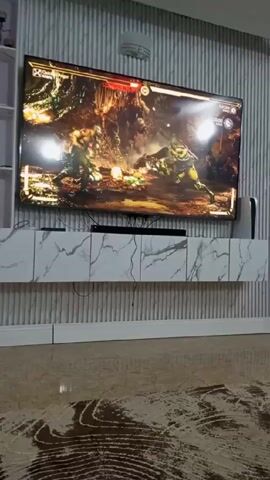 The video shows a wide shot of a living room with a large television mounted on a wall. The TV is displaying the video game Mortal Kombat, with two characters engaged in a fight. A white console, likely a PlayStation 5, is visible to the right of the TV. Below the TV is a white marble-patterned cabinet. The wall behind the TV has a textured, vertically ribbed design. The floor is covered with a patterned carpet. An audible sound like a child or baby crying can be heard in the background.