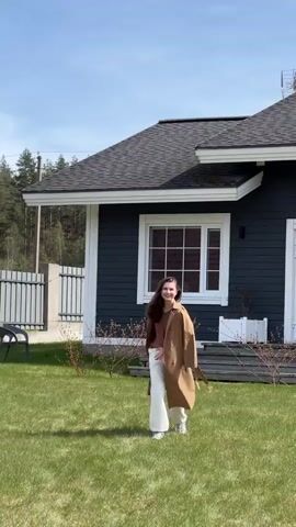 A young woman with long brown hair is running towards the camera on a green lawn. She is wearing a brown V-neck shirt, cream-colored pants, and a tan trench coat. She smiles brightly as she runs. Behind her is a dark grey house with white trim and a grey roof. There are bare shrubs planted in front of the house. A wooden fence is visible on the left side of the frame, and trees can be seen in the distance. The sky is blue with some clouds.