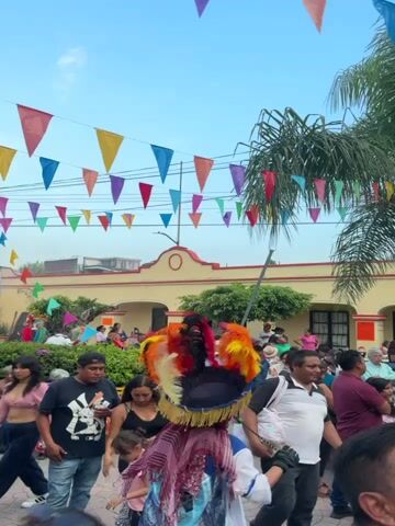 The video shows a crowded outdoor event in Mexico. The camera is positioned low to the ground, offering an eye-level view of the bustling scene. The crowd is a mix of people of different ages, with some sitting and others standing. There are a few palm trees scattered throughout the scene and colorful triangle banners hanging overhead, adding to the festive atmosphere. A man wearing a vibrant headdress made of feathers stands in the foreground, adding to the cultural richness of the event. In the background, there is a stage covered with a green tarp, possibly set up for live music or performances. There's a building in the background and a street light on the side. The video seems to capture a community celebration or cultural gathering.