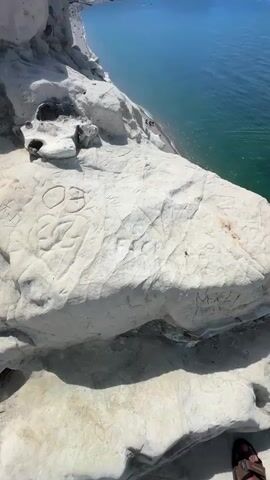 The video pans from a close-up of a textured white rock formation with carvings to a wider view of a beach and the ocean. The rock is large and weathered, with visible indentations and lines that appear to be deliberate markings or graffiti. In the background, a long, pebble beach stretches along a turquoise ocean. The water is clear, with visible green and blue hues. Several small figures, presumably people, are scattered on the beach, some lying down and others walking. Further out in the ocean, a few rocky outcrops rise from the water, and a distant coastline with cliffs is visible. The sky is a clear, bright blue with a few faint clouds. The sun is shining, casting shadows on the rock in the foreground. The overall impression is a scenic coastal landscape on a sunny day.