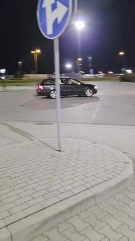 A black car is seen turning in a parking lot at night. The car appears to be a sedan with tinted windows. In the background, several parked trucks and a bus are visible. The lighting in the scene is from streetlights.