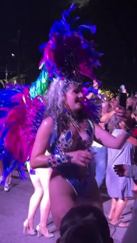 A woman in a vibrant, feathered costume, adorned with blue and pink plumes and a jeweled headdress, dances in the street at night. Her costume features a blue, sequined leotard with jeweled embellishments on the neckline and hips. She holds a phone in her right hand, seemingly recording herself or the event. Her expression is joyful and her mouth is open as if singing or laughing. Behind her, a crowd of people watch the performance, some holding up their own phones to capture the moment. The environment appears to be an outdoor street festival or parade, with some string lights visible in the background and a dark, nighttime sky. The lighting is atmospheric, with spotlights illuminating the dancer. The crowd consists of individuals of various ages and ethnicities, dressed in casual attire. The dancer's movements are lively and energetic, suggesting a celebratory atmosphere.