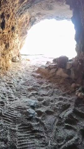 A first-person perspective video captures a sandy path leading out of a cave to a bright, sunny beach. The cave walls are rough and textured, with visible footprints in the sand on the path. Rocks and pebbles are scattered on the ground. The exit of the cave opens to reveal the ocean with clear blue water, rocky shorelines, and a bright sky. Trees are visible in the distance on the right side of the frame. The video pans slightly, offering a wider view of the beach and sea.