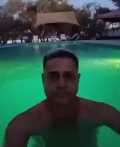A man is shown taking a selfie video in a swimming pool. The water's color changes from green to blue and purple during the recording, indicating the pool's lighting system is rotating the hue. The man is shirtless and looks directly into the camera. Behind him, there are some lounge chairs and a thatched-roof structure. The video is taken at night.