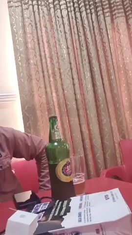 A man is sitting at a red table, wearing a pink button-down shirt. He is looking at the camera with a neutral expression, and his right hand is on his forehead.  There is a green beer bottle and a glass of beer on the table, along with a wallet, a lighter, a pack of cigarettes, and a water bottle.  The background consists of patterned curtains and a white wall.