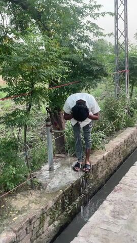 The video captures scenes from a rural setting, likely in Karamsar, Pakistan.

The first segment shows a young man, dressed in a white shirt, dark trousers, and black sandals, using a hand pump. He bends over the pump, washing his face and hands as water gushes out. The pump is positioned next to a narrow concrete channel or drain, filled with dark water. In the background, there are lush green trees, unpaved ground, and a metal utility pole, indicating an outdoor, possibly agricultural or village environment. The ground around the pump is wet from the splashing water.

The camera then quickly pans across a dusty, unpaved area with blurred outlines of distant buildings and foliage, suggesting a transition within the locality.

The final segment focuses on the entrance of a house or dwelling. The structure features brick walls, some with exposed brick and others painted. A blue metal gate is visible, behind which a buffalo and a smaller dark animal can be seen. On a raised porch-like area near the house, a red wooden stool supports what appears to be a part of a hand-operated machine. The ground in this area is also unpaved.

The overall impression is that of everyday life in a Pakistani village. The weather seems to be daytime, with a somewhat overcast or hazy sky, providing diffused light. No visible text is present in the video. The man's actions suggest a routine activity, without any strong emotional display.