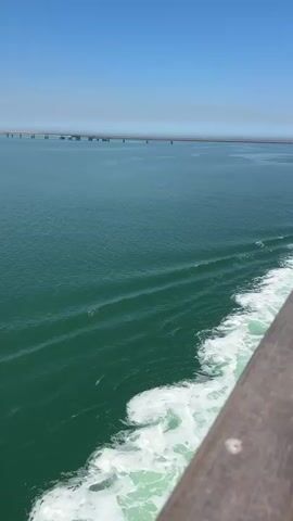 The video shows a view from the side of a moving boat. The water is a deep green, and the boat is creating white foam as it moves. In the distance, a long bridge stretches across the water. The sky is clear and blue.