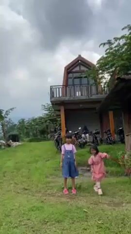 Two young girls are on a grassy slope near a wooden structure. One girl is wearing a pink dress and the other a blue denim overall dress. They run across the grass, laughing and smiling.  Behind them, parked motorcycles are visible near a two-story building with a balcony.  In the background, there are terraces and a cloudy sky. The girls run towards a gazebo-like structure where people are seated and socializing.