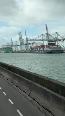 The video depicts a port scene under a cloudy sky. A large cargo ship, emblazoned with "CMA CGM," sits in the water, dwarfed by numerous gantry cranes lining the dock. Another cargo ship is visible to the left, green in color. A concrete barrier runs along the edge of the road, marked with white lane lines, from which the footage is shot. The water is choppy with small waves.