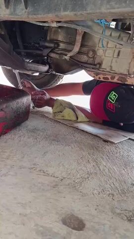 A person wearing a cap and a yellow cloth is lying on the ground underneath a vehicle. They are using a tool to work on the underside of the vehicle, and a red container is positioned to catch any drips. The ground appears to be concrete.