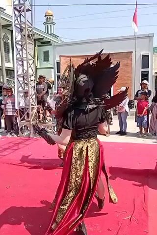 This video shows a performance with several individuals in elaborate costumes. The primary focus is on two performers in the foreground, wearing dark and red elaborate outfits with large, wing-like headpieces and flowing skirts. They are on a red carpet, and appear to be dancing or posing. In the background, there are spectators of various ages, as well as buildings and a stage structure. The overall environment appears to be an outdoor event.