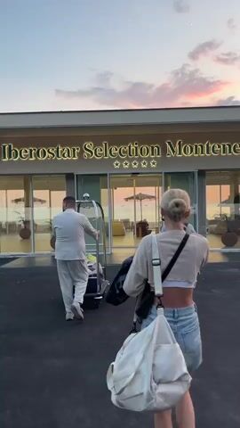 This media file depicts a twilight scene outside the Iberostar Selection Montenegro hotel in Mišići, Montenegro. Three individuals are visible, two women and a hotel staff member. The two women are walking towards the hotel entrance, one in the foreground carrying a large white bag and a black purse, wearing a cropped beige top and denim shorts, with her blonde hair pulled up in a bun. The second woman, further ahead, wears a grey t-shirt and shorts, with a blue patterned backpack. A male hotel employee, dressed in a light-colored uniform, is pushing a luggage cart filled with bags towards the entrance, walking ahead of the women.

The hotel entrance is prominent, with large glass doors and windows revealing a brightly lit interior featuring lounge furniture and glimpses of the sea beyond. The "Iberostar Selection Montenegro" signage is illuminated with golden lights, accompanied by five stars. The sky is painted with soft hues of pink, orange, and purple, indicating either sunrise or sunset, with a crescent moon visible. The overall atmosphere is serene and suggests the arrival or departure of guests at a resort.