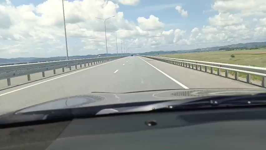 The video shows a first-person perspective from a car driving on a highway. The road is paved and has white lane markings. Guardrails are visible on both sides of the road. In the distance, there are hills and a cloudy sky. Streetlights are spaced along the highway. The dashboard of the car is visible in the foreground.