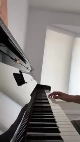 The video shows a low-angle view of a person playing a black piano. The person's hands are visible on the keys. The piano is positioned against a white wall near a window covered by blinds. A painting is partially visible on the wall. The camera remains still throughout the video.