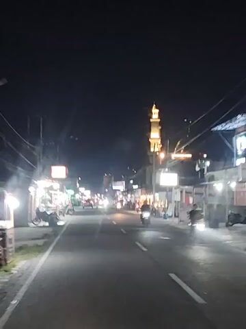 The video shows a street scene at night. The street is paved with white lines indicating lanes. On both sides of the street, there are buildings and stalls, many of which are illuminated by lights. There are several motorcycles and a few cars visible on the street, some of which appear to be moving. In the distance, a tall, slender tower, possibly a minaret of a mosque, is illuminated with yellow lights. The sky is dark, indicating nighttime. The overall impression is that of a busy street in a town or city at night.