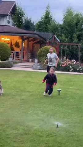 A father and son are running on a grassy lawn. The father is in the background, wearing a t-shirt and shorts, and the son is in the foreground, wearing a jacket and pants. A sprinkler is spraying water on the lawn, and a rose bush is visible in the background. The scene appears to be in a backyard, with a house in the background.