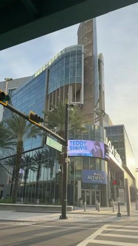 The video shows an exterior view of a large modern building with glass windows and a spiraling architectural feature at the top. In the foreground, there is a street with a crosswalk and traffic lights. A street sign indicates "Church St." Palm trees are visible along the sidewalk. A large digital billboard on the building displays an advertisement for "TEDDY SWIMS" and below it, another sign reads "MAGIC AUTHENTICS". The sky is bright, suggesting daylight. A few people are walking on the sidewalk in the distance. The camera appears to be moving slightly, possibly from a vehicle.