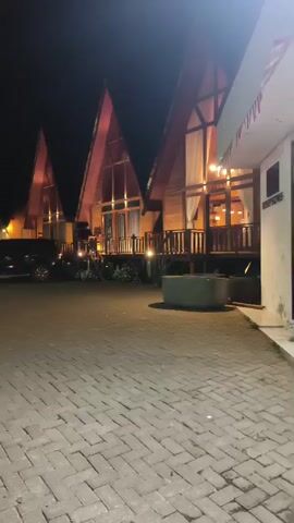 A nighttime shot captures a row of A-frame wooden cabins with illuminated windows, suggesting an accommodation setting. The cabins are built on stilts, with wooden decks and railings. The foreground is paved with interlocking bricks, and a dark-colored SUV is parked to the left. Further back, a teal pickup truck is visible. To the right, a modern building with glass doors and windows is also lit up. A prominent illuminated signpost is present, displaying information about facilities such as parking, Wi-Fi, and a cafe. The overall scene depicts a tranquil, possibly resort-like environment at night.