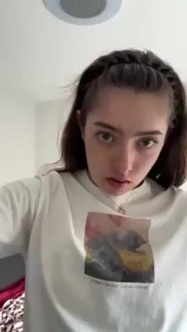 A young woman stands in a brightly lit room, wearing a white t-shirt with a graphic print. She has fair skin, dark hair, and green eyes. The woman is adjusting a dark headband with braided detailing on her head. The camera remains still at a low angle. The room's decor is simple, with a light-colored wall and a ceiling light visible. A patterned fabric is briefly seen in the lower left corner. The woman looks directly at the camera with a slight frown before adjusting her hair and looking over her shoulder. The print on the back of her shirt is partially visible, featuring an image of a person on a beach.