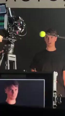The video shows a man in a black t-shirt, who appears to be an actor, standing in front of a black backdrop. He is grimacing, as though angry. There is a boom mic over his head. A professional video camera is positioned on a tripod in front of him, with someone adjusting it. The camera is also pointed at him. The monitor for the camera shows the same view, so it looks as if the man is watching himself make this expression.