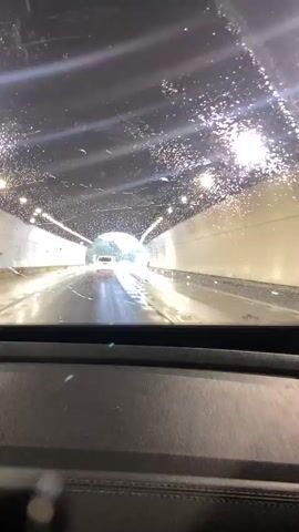 The video is a first-person view from inside a car driving through a tunnel. The windshield is wet with raindrops, distorting the view. The tunnel is illuminated by a series of lights along the ceiling and walls. The road ahead is wet and reflective, suggesting it is raining outside. There are other cars in the tunnel, including one in front that is visible with its taillights illuminated. As the car progresses, it emerges from the tunnel into an overcast, rainy environment with trees and road signs visible.