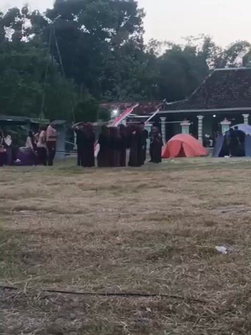The video shows an outdoor scene, likely a campsite, during daylight hours. Several tents are pitched on a grassy field. In the background, there are traditional-looking buildings with tiled roofs. A group of people, some wearing dark uniforms, are standing in formation, appearing to be saluting or in a formal pose. The overall atmosphere suggests some kind of organized activity or event taking place outdoors.