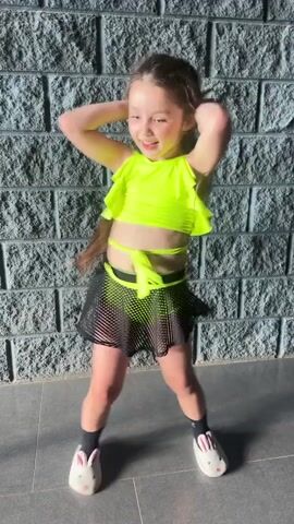 A young girl with long brown hair dances in front of a grey brick wall. She is wearing a bright neon yellow halter-top crop top with ruffled detailing, a matching neon yellow sash tied around her waist, and a sheer black mesh skirt with a neon yellow lining. She also wears black socks and white fuzzy slippers with bunny ears. Her hair is long and flowing as she moves. The girl appears happy and energetic, with a wide smile and her tongue sticking out in one shot. The scene is outdoors, with the sun casting shadows on the ground, suggesting it is daytime. The ground is paved with large, grey tiles. There is no visible text in the media file. The location is Sverdlovskiy, Russia.