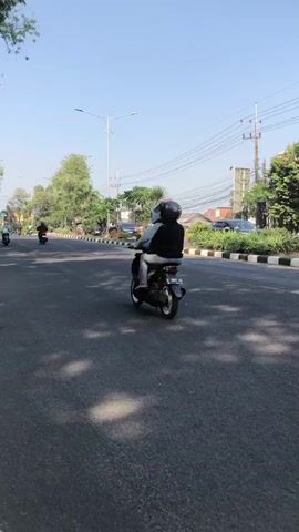 A vertical video shows a street scene with multiple motorcycles. In the foreground, a motorcycle with a dark-colored cart attached to the rear is moving away from the camera. The cart appears to be filled with black trash bags. The rider is wearing a black helmet and a black t-shirt. In the middle ground, another motorcycle is also moving away from the camera. The rider is wearing a black helmet and a black jacket. Further in the background, more motorcycles are visible on the road. The environment appears to be a sunny day with trees lining the street and overhead power lines. The road is paved and has lane markings. There is a sidewalk on the left side of the road. A silver SUV is also visible on the right side of the road.