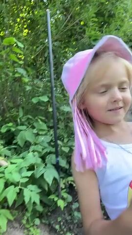 The video features a young girl with blonde hair, wearing a pink bucket hat and a white tank top with black patterned leggings. She is standing in a lush green outdoor setting, likely a garden or wooded area, with a rough wooden post supporting two pieces of clothing on either side. Behind her, a wooden sign with Cyrillic text and a fish graphic is visible. The girl is looking towards the camera with a playful expression and appears to be holding something in her hands. The overall atmosphere suggests a casual, outdoor activity.

The visible text on the sign appears to be "Я всё вижу" which translates to "I see everything" in Russian. The lighting suggests it is daytime. The environment is dense with foliage, indicating a natural or semi-cultivated area. The presence of drying clothes on a makeshift rack and the girl's attire suggest a possibly warm weather and an informal setting. The girl's expression conveys a sense of fun and engagement.