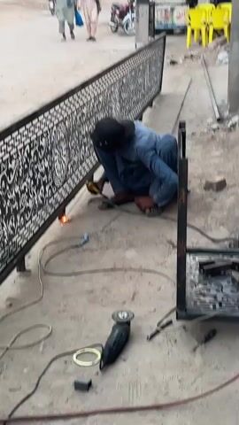 An outdoor scene captures a welder performing arc welding on an elaborate metal balustrade or fence. The welder, wearing a dark hood/cap and light blue attire, crouches on a concrete-like surface, generating bright sparks from the welding arc. The balustrade is finished in black with intricate silver/white patterns, featuring a repeating Greek key border, floral scrollwork, and circular medallions. Welding cables and an angle grinder are visible on the ground next to the metalwork. In the background, two individuals walk down what appears to be a street, and a motorcycle is parked. Further in the distance, bright yellow chairs are stacked, possibly indicating an eatery. The activity takes place during daytime under bright conditions, suggesting a workshop or street-side fabrication in Nasheman Colony, Pakistan. No discernible text is visible.