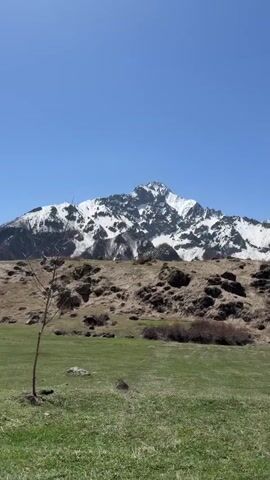 The video presents a serene landscape featuring a verdant field leading to a rugged, rocky hill. A majestic snow-capped mountain dominates the background under a clear, blue sky. The wind is audible.