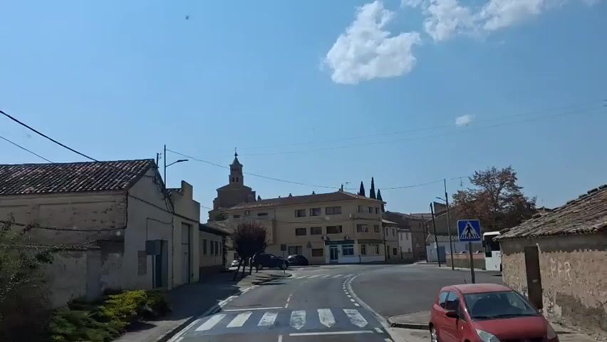 The video captures a view of a street in what appears to be a small town. The scene is dominated by a clear blue sky with a few scattered clouds. In the foreground, there is a road with a pedestrian crossing. On the left side of the road, there is a low, light-colored building with a tiled roof and what looks like a garage door. Further down the street, another building with a similar roof is visible. Across the street, a larger, multi-story building with a cream-colored facade and numerous windows is present. To the right of this building, a prominent church tower with a cross on top rises against the sky. There are also some trees, including one with red foliage, and power lines crisscrossing the frame. A white bus is parked on the right side of the road, and a red car is partially visible in the lower right corner. There are no visible people in the immediate vicinity, though the presence of cars and a bus suggests it is a populated area. The overall impression is a sunny day in a European town.