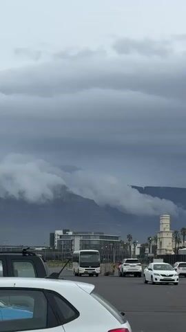 The video shows an overcast day in what appears to be a coastal urban area. In the background is a mountain partially obscured by low-hanging clouds. Several vehicles are visible in the foreground, including cars, a bus, and what may be a tour bus. There are buildings, palm trees, and a clock tower in the middle ground. The overall impression is one of a city near a mountain range on a cloudy day.