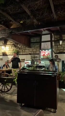 A man in a black t-shirt and khaki shorts stands at a microphone, speaking. To his right, two individuals, a man in a white shirt and a woman in a denim jacket, sit behind a table with musical equipment. The scene appears to be an outdoor restaurant or venue with stone walls and a wooden ceiling covered in vines. A decorative cart is positioned to the left of the stage. The lighting changes from purple to green and back to blue throughout the clip.