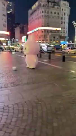 The video shows a person in a large, pink rabbit costume standing on a cobblestone street at night. The person in the costume is facing the camera with a smiling expression. Behind the rabbit costume to the left is a person in a short skirt. There are buildings and traffic in the background. A small white box is on the ground in front of the costume.