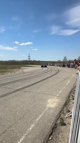 The video shows a green sports car drift racing on an asphalt track, with a crowd of spectators lining the edge. The car is decorated with stickers and has white rims. There are visible tire marks on the road. In the background are power lines, trees, and a clear blue sky with scattered clouds. A person in a white hooded shirt and pants stands on the track waving a white paper, followed by another person wearing a black jacket and pants, appearing to be signaling or directing the car. The camera is positioned on the side of the track, and the angle captures the car as it passes.