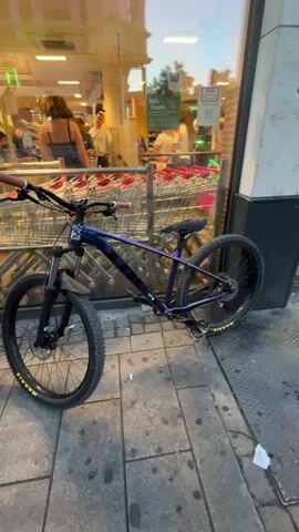 The video shows two mountain bikes parked outside a storefront with large glass windows. The bikes are parked on a paved sidewalk with a metal grate to the right. Inside the store, people and shopping carts are visible. The lighting suggests it is late afternoon or early evening. The first bike, on the left, is dark purple and black with brown handlebars. The second bike, on the right, is predominantly black with white accents on the wheels and frame. The camera pans slowly from the first bike to the second.