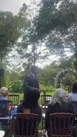 The video shows a wedding ceremony taking place in a garden or wooded area. There are rows of chairs set up on a lawn, facing a floral archway that serves as the backdrop for the ceremony. Guests are seated, facing forward. A man in a dark suit stands at the front, addressing the audience with a microphone. There is movement in the background as other individuals in formal attire approach the front. The environment appears to be lush with trees, greenery, and possibly some lighting strung in the branches.